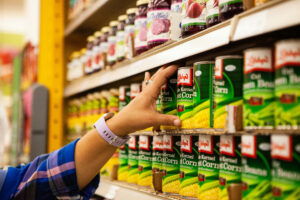 A woman's hand reaches for a can of corn at the grocery store.
