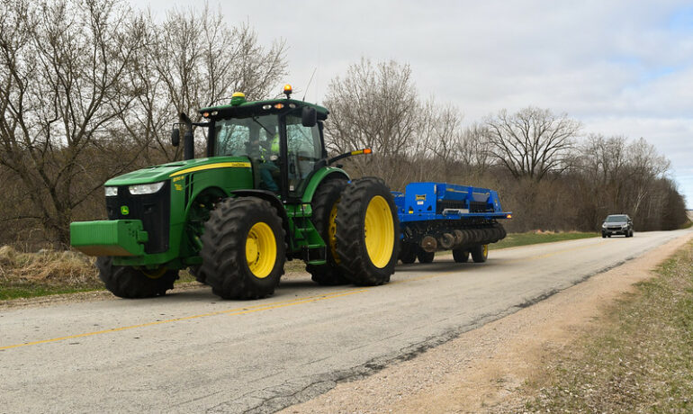 A tractor pulling a disk is driving on a road followed by a Chevrolet Traverse car.